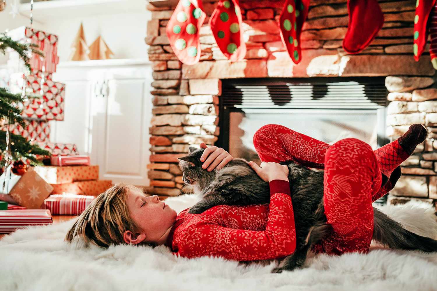 Junge liegt auf einem weißen, flauschigen Teppich mit einer Katze auf der Brust neben einem Weihnachtsbaum